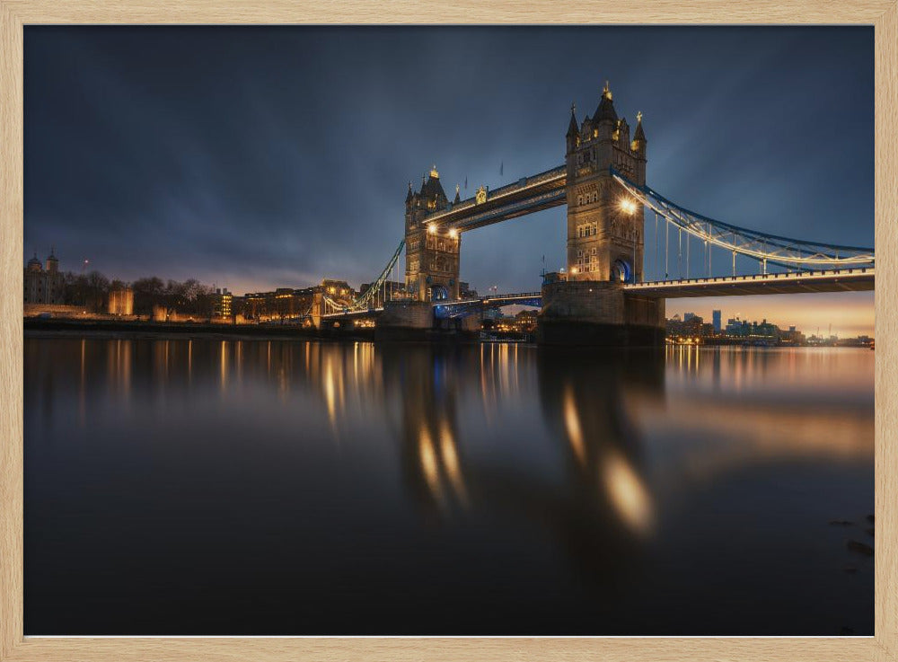 A long-exposure photograph of London's Tower Bridge at dusk, brilliantly illuminated with golden lights that cast shimmering reflections on the calm River Thames. The sky is a deep, dark blue, and the city skyline glows in the background, all enclosed within a silver frame. Print