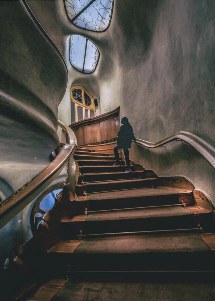 A low-angle view up a dark, winding wooden staircase inside a building with surreal, organic architecture. A person in a dark coat is walking up the stairs towards uniquely shaped windows that show a blue sky, contrasting with the dimly lit, textured gray walls. Print