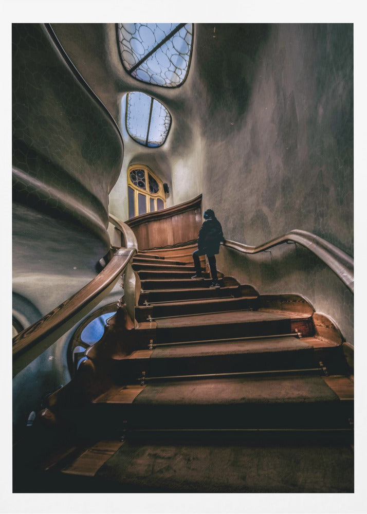 A low-angle view up a dark, winding wooden staircase inside a building with surreal, organic architecture. A person in a dark coat is walking up the stairs towards uniquely shaped windows that show a blue sky, contrasting with the dimly lit, textured gray walls. Print