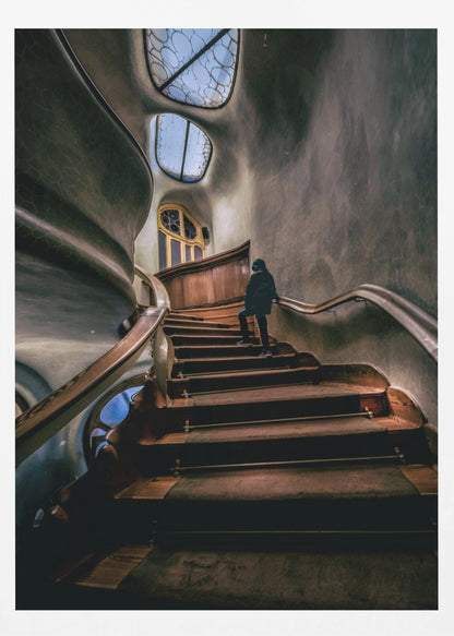 A low-angle view up a dark, winding wooden staircase inside a building with surreal, organic architecture. A person in a dark coat is walking up the stairs towards uniquely shaped windows that show a blue sky, contrasting with the dimly lit, textured gray walls. Print