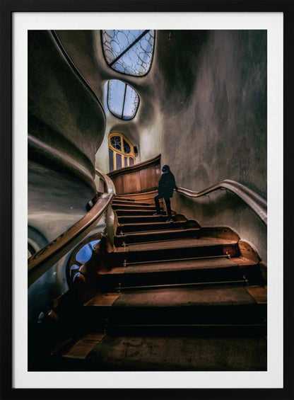 A low-angle view up a dark, winding wooden staircase inside a building with surreal, organic architecture. A person in a dark coat is walking up the stairs towards uniquely shaped windows that show a blue sky, contrasting with the dimly lit, textured gray walls. Print