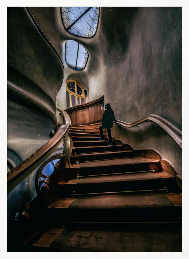 A low-angle view up a dark, winding wooden staircase inside a building with surreal, organic architecture. A person in a dark coat is walking up the stairs towards uniquely shaped windows that show a blue sky, contrasting with the dimly lit, textured gray walls. Print