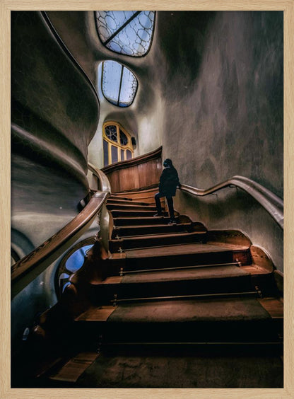 A low-angle view up a dark, winding wooden staircase inside a building with surreal, organic architecture. A person in a dark coat is walking up the stairs towards uniquely shaped windows that show a blue sky, contrasting with the dimly lit, textured gray walls. Print