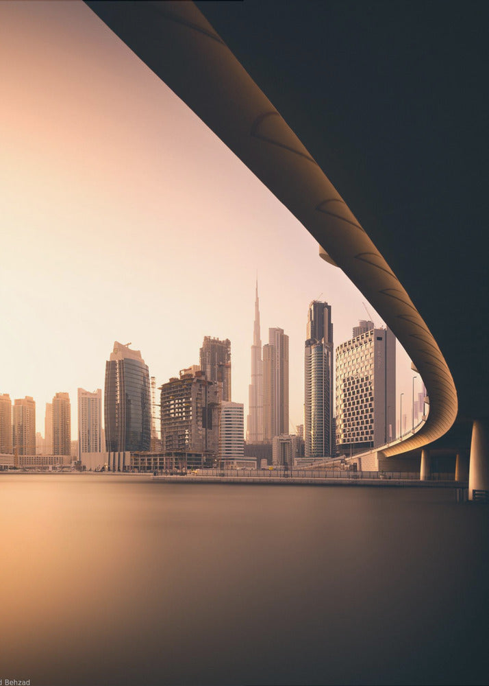 A view of the Dubai skyline, including the Burj Khalifa, framed by the sweeping curve of a bridge in the foreground. The city is bathed in the warm, soft light of a sunrise or sunset, with its reflection visible on the calm water below. Decor