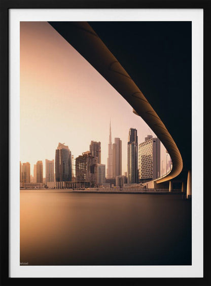 A view of the Dubai skyline, including the Burj Khalifa, framed by the sweeping curve of a bridge in the foreground. The city is bathed in the warm, soft light of a sunrise or sunset, with its reflection visible on the calm water below. Decor