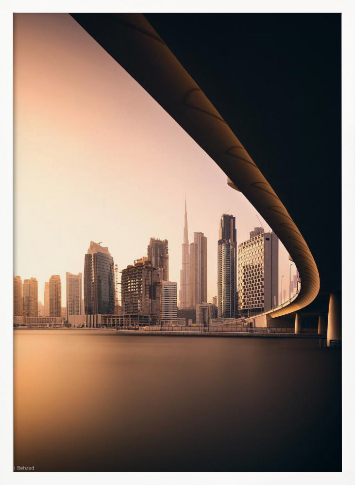 A view of the Dubai skyline, including the Burj Khalifa, framed by the sweeping curve of a bridge in the foreground. The city is bathed in the warm, soft light of a sunrise or sunset, with its reflection visible on the calm water below. Decor