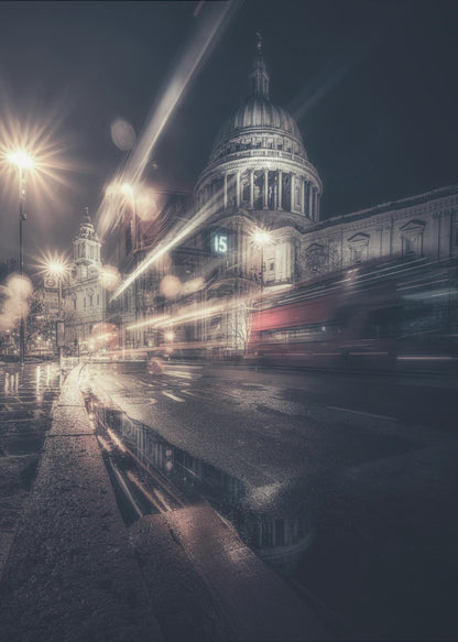 A dynamic long-exposure photograph of a wet London street at night, with the iconic dome of St. Paul's Cathedral illuminated in the background. Streaks of light from passing traffic, including a red bus, blur across the scene, while the glistening pavement reflects the cathedral and bright streetlights. Decor