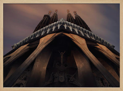 A dramatic, low-angle shot of the Sagrada Familia cathedral, focusing on the intricate details of the Passion Facade against a dusky purple and orange sky. The spires tower above, while deep shadows highlight the unique, angled columns and the central sculpture of the crucifixion. Poster