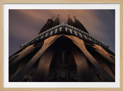 A dramatic, low-angle shot of the Sagrada Familia cathedral, focusing on the intricate details of the Passion Facade against a dusky purple and orange sky. The spires tower above, while deep shadows highlight the unique, angled columns and the central sculpture of the crucifixion. Poster