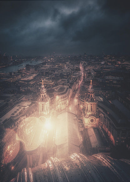 A dramatic, high-angle nighttime photograph of a sprawling city, likely London, viewed from the top of a cathedral. The foreground shows the curved dome, while below, two towers and the long roof of the church are brightly illuminated. The city stretches into the distance with countless golden lights under a dark, stormy, and cloud-filled sky. Poster