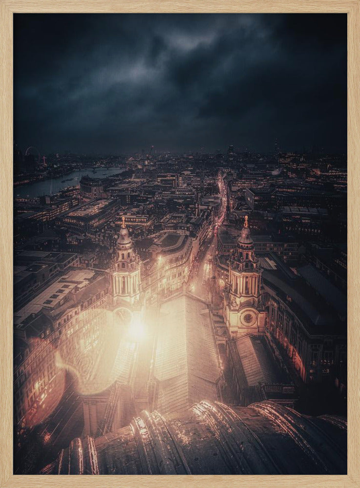 A dramatic, high-angle nighttime photograph of a sprawling city, likely London, viewed from the top of a cathedral. The foreground shows the curved dome, while below, two towers and the long roof of the church are brightly illuminated. The city stretches into the distance with countless golden lights under a dark, stormy, and cloud-filled sky. Poster
