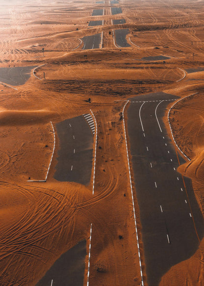 An aerial view of a multi-lane, black asphalt road being consumed by vast, rolling sand dunes of a deep orange color. The sand covers sections of the road, creating a surreal pattern of man-made lines and natural, windswept textures. Print