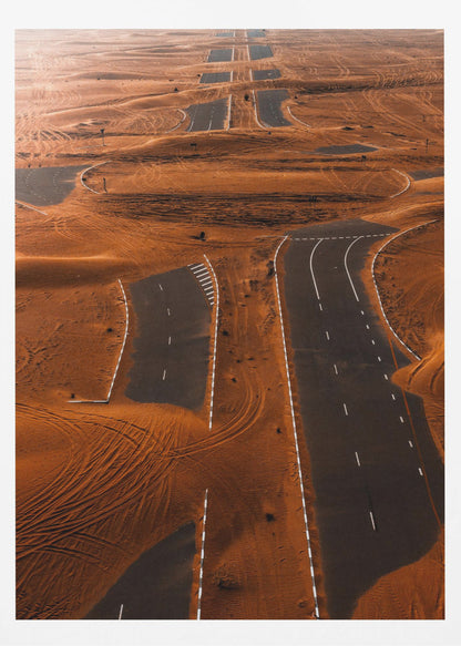 An aerial view of a multi-lane, black asphalt road being consumed by vast, rolling sand dunes of a deep orange color. The sand covers sections of the road, creating a surreal pattern of man-made lines and natural, windswept textures. Print