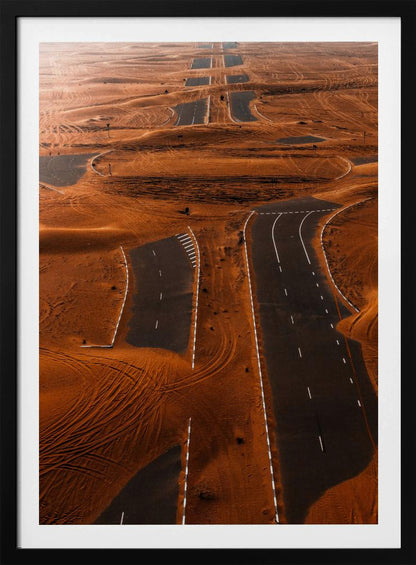 An aerial view of a multi-lane, black asphalt road being consumed by vast, rolling sand dunes of a deep orange color. The sand covers sections of the road, creating a surreal pattern of man-made lines and natural, windswept textures. Print
