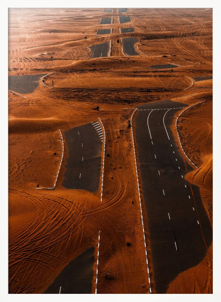 An aerial view of a multi-lane, black asphalt road being consumed by vast, rolling sand dunes of a deep orange color. The sand covers sections of the road, creating a surreal pattern of man-made lines and natural, windswept textures. Print