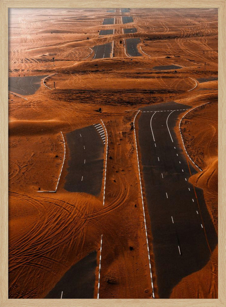 An aerial view of a multi-lane, black asphalt road being consumed by vast, rolling sand dunes of a deep orange color. The sand covers sections of the road, creating a surreal pattern of man-made lines and natural, windswept textures. Print