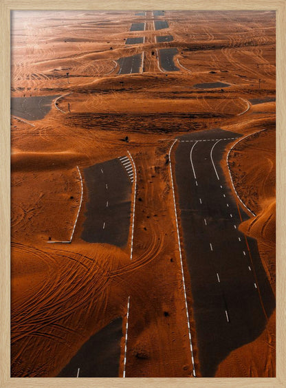An aerial view of a multi-lane, black asphalt road being consumed by vast, rolling sand dunes of a deep orange color. The sand covers sections of the road, creating a surreal pattern of man-made lines and natural, windswept textures. Print