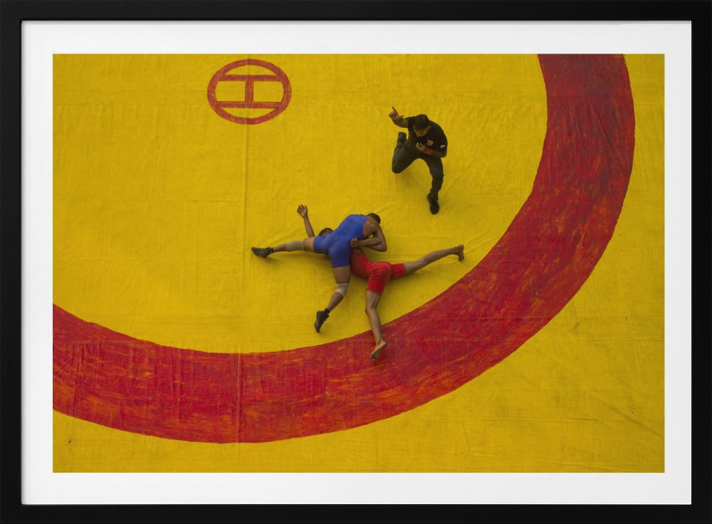 An aerial, high-angle photograph of a wrestling match in a silver frame. Two wrestlers, one in a blue singlet and the other in red, are locked in a hold on a vibrant yellow mat marked with a thick, painted red circle. A referee dressed in black stands nearby, gesturing. Artwork
