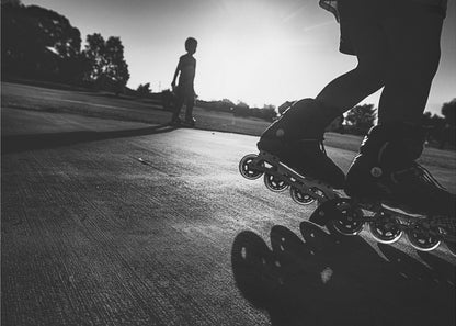 A low-angle, black and white photograph of two people rollerblading on a paved path at sunset. The foreground is a close-up of one person's rollerblades and their long shadow, while the second person is silhouetted in the distance against the bright sun. Artwork