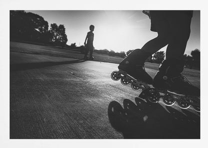 A low-angle, black and white photograph of two people rollerblading on a paved path at sunset. The foreground is a close-up of one person's rollerblades and their long shadow, while the second person is silhouetted in the distance against the bright sun. Artwork