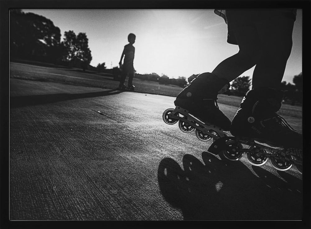 A low-angle, black and white photograph of two people rollerblading on a paved path at sunset. The foreground is a close-up of one person's rollerblades and their long shadow, while the second person is silhouetted in the distance against the bright sun. Artwork