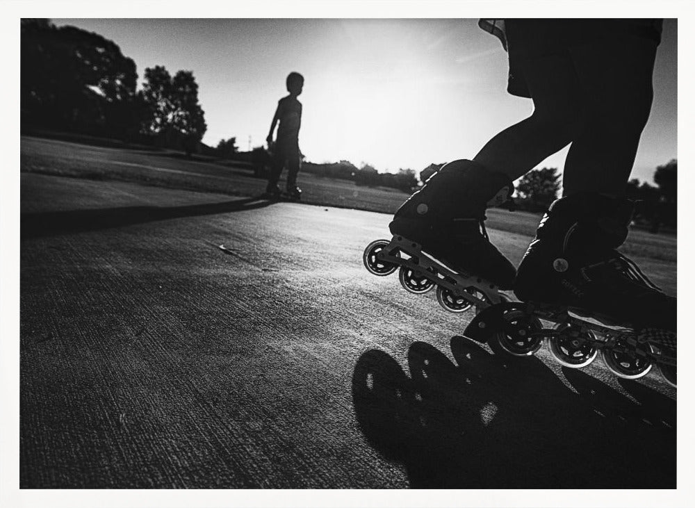 A low-angle, black and white photograph of two people rollerblading on a paved path at sunset. The foreground is a close-up of one person's rollerblades and their long shadow, while the second person is silhouetted in the distance against the bright sun. Artwork