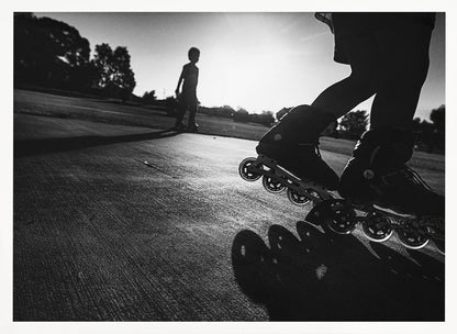 A low-angle, black and white photograph of two people rollerblading on a paved path at sunset. The foreground is a close-up of one person's rollerblades and their long shadow, while the second person is silhouetted in the distance against the bright sun. Artwork