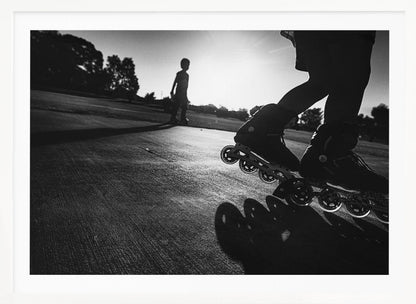A low-angle, black and white photograph of two people rollerblading on a paved path at sunset. The foreground is a close-up of one person's rollerblades and their long shadow, while the second person is silhouetted in the distance against the bright sun. Artwork