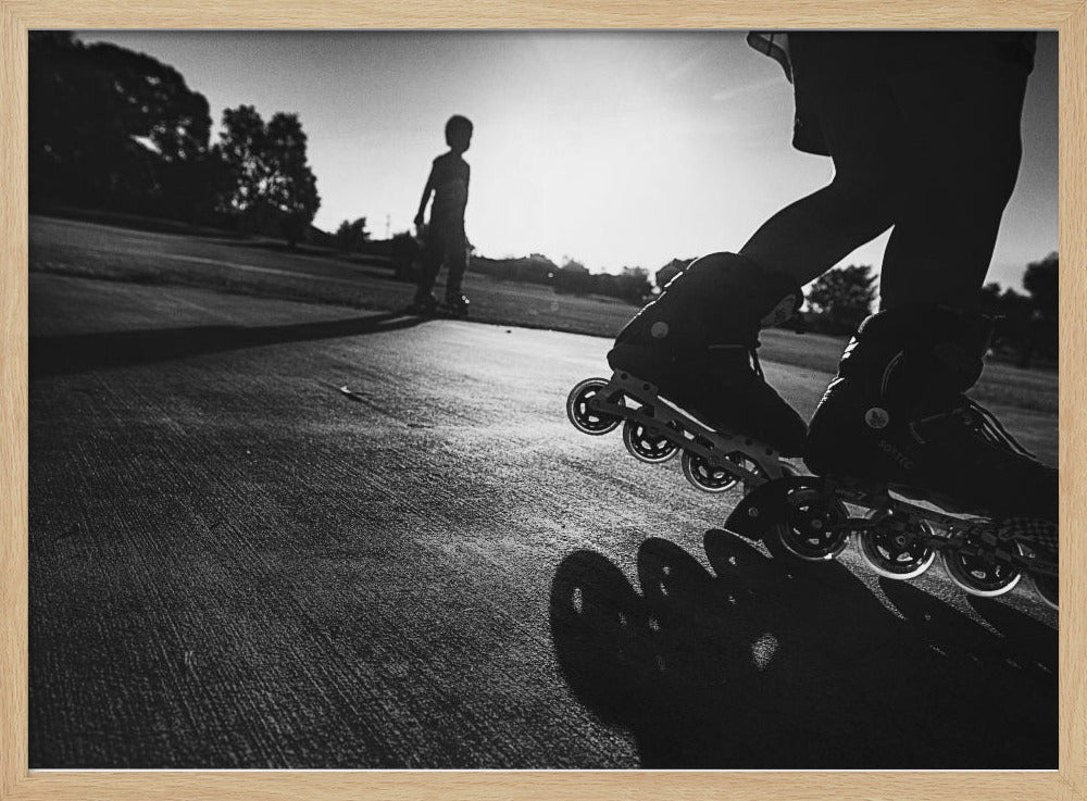 A low-angle, black and white photograph of two people rollerblading on a paved path at sunset. The foreground is a close-up of one person's rollerblades and their long shadow, while the second person is silhouetted in the distance against the bright sun. Artwork
