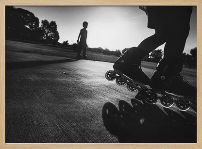 A low-angle, black and white photograph of two people rollerblading on a paved path at sunset. The foreground is a close-up of one person's rollerblades and their long shadow, while the second person is silhouetted in the distance against the bright sun. Artwork