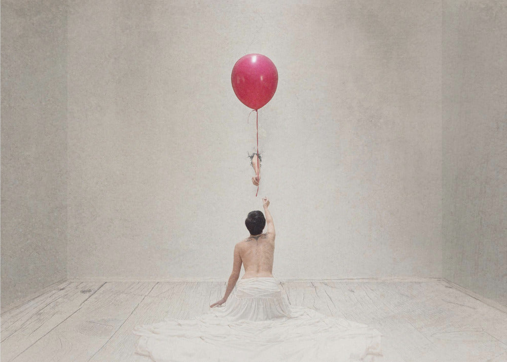 A surreal fine art photograph of a person with a bare back and a long white skirt, sitting on the floor of a minimalist grey room. They are reaching up towards a small figure hanging from the string of a large, bright red balloon that floats towards the ceiling. The image is enclosed in a silver frame. Artwork