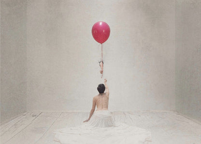 A surreal fine art photograph of a person with a bare back and a long white skirt, sitting on the floor of a minimalist grey room. They are reaching up towards a small figure hanging from the string of a large, bright red balloon that floats towards the ceiling. The image is enclosed in a silver frame. Artwork
