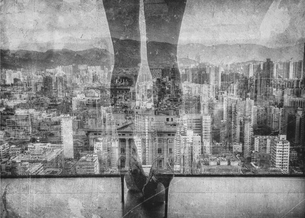 A framed, black and white artistic photograph featuring a double exposure. A vast, dense cityscape with distant mountains is visible through the translucent silhouette of a person's legs standing on a balcony edge. Poster
