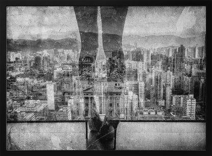 A framed, black and white artistic photograph featuring a double exposure. A vast, dense cityscape with distant mountains is visible through the translucent silhouette of a person's legs standing on a balcony edge. Poster
