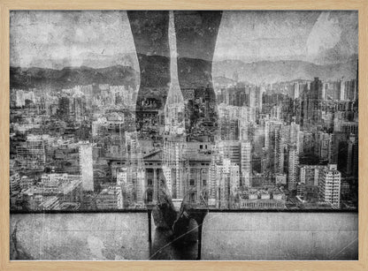 A framed, black and white artistic photograph featuring a double exposure. A vast, dense cityscape with distant mountains is visible through the translucent silhouette of a person's legs standing on a balcony edge. Poster