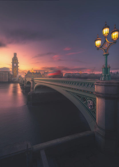 A breathtaking twilight view of Westminster Bridge in London, with the iconic Big Ben illuminated in the background against a purple and orange sunset sky. A vintage street lamp glows warmly in the foreground, and the motion blur of a red bus crosses the bridge over the calm River Thames. Print