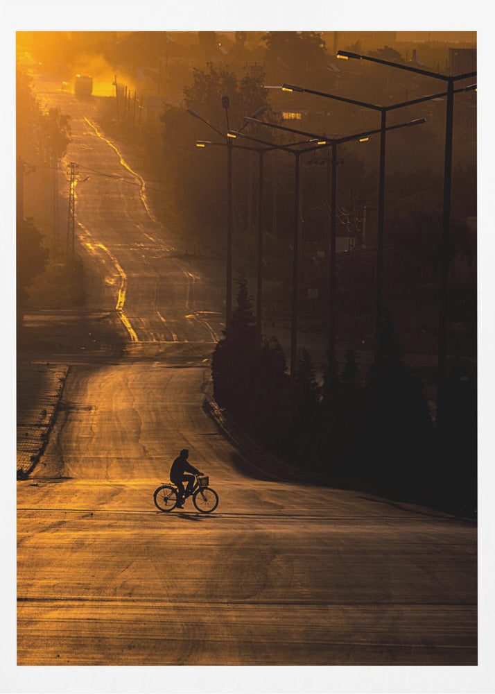 A silhouette of a person riding a bicycle across a wide, winding road illuminated by the warm, golden light of a sunrise or sunset. The road stretches uphill into the distance, and tall streetlights stand in shadow on the right. Artwork