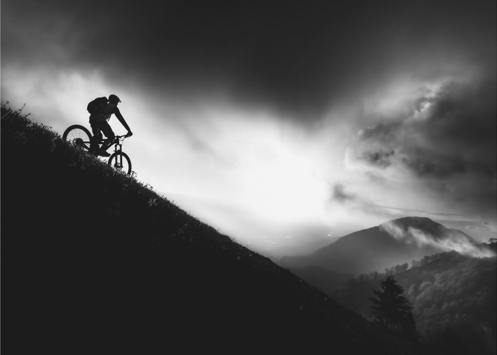 A striking black and white photo of a mountain biker's silhouette on a steep decline, set against a dramatic, light-filled cloudy sky with distant, misty mountains. Artwork