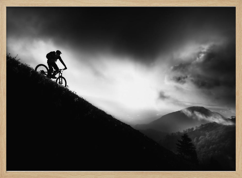 A striking black and white photo of a mountain biker's silhouette on a steep decline, set against a dramatic, light-filled cloudy sky with distant, misty mountains. Artwork