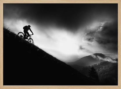 A striking black and white photo of a mountain biker's silhouette on a steep decline, set against a dramatic, light-filled cloudy sky with distant, misty mountains. Artwork
