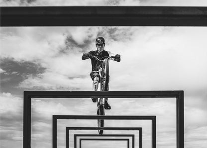A striking black and white photograph taken from a low angle, showing a cyclist in full gear balancing on a mountain bike. The cyclist is positioned centrally above a series of black rectangular frames that create a receding, tunnel-like visual effect against a cloudy sky. The entire image is presented within a silver picture frame. Artwork