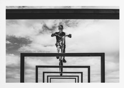 A striking black and white photograph taken from a low angle, showing a cyclist in full gear balancing on a mountain bike. The cyclist is positioned centrally above a series of black rectangular frames that create a receding, tunnel-like visual effect against a cloudy sky. The entire image is presented within a silver picture frame. Artwork