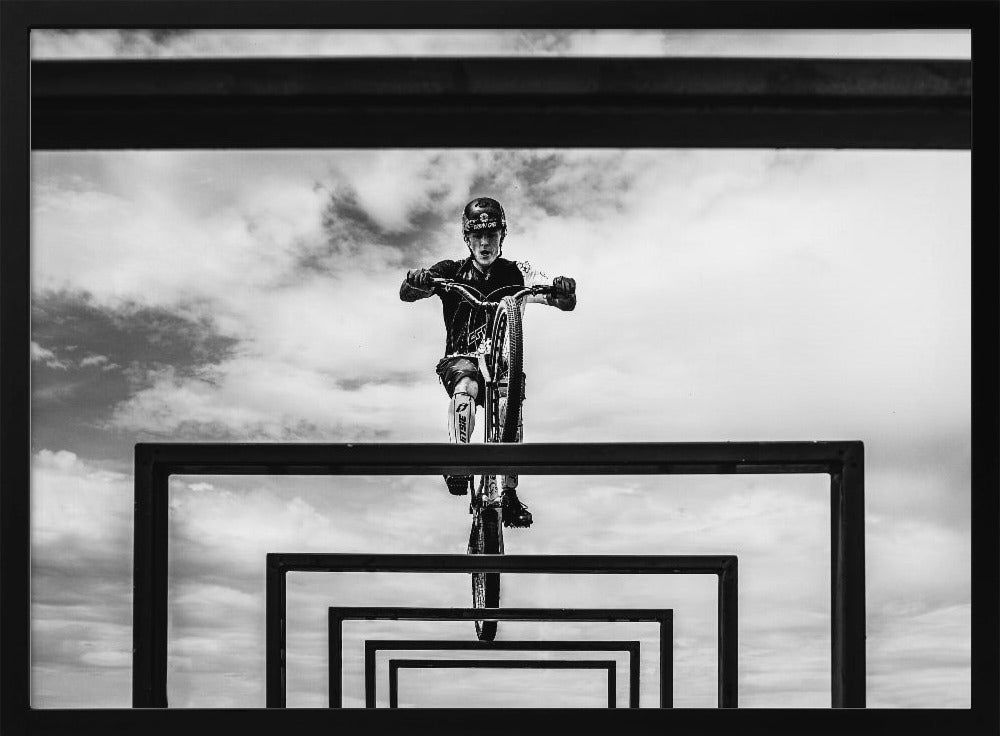 A striking black and white photograph taken from a low angle, showing a cyclist in full gear balancing on a mountain bike. The cyclist is positioned centrally above a series of black rectangular frames that create a receding, tunnel-like visual effect against a cloudy sky. The entire image is presented within a silver picture frame. Artwork