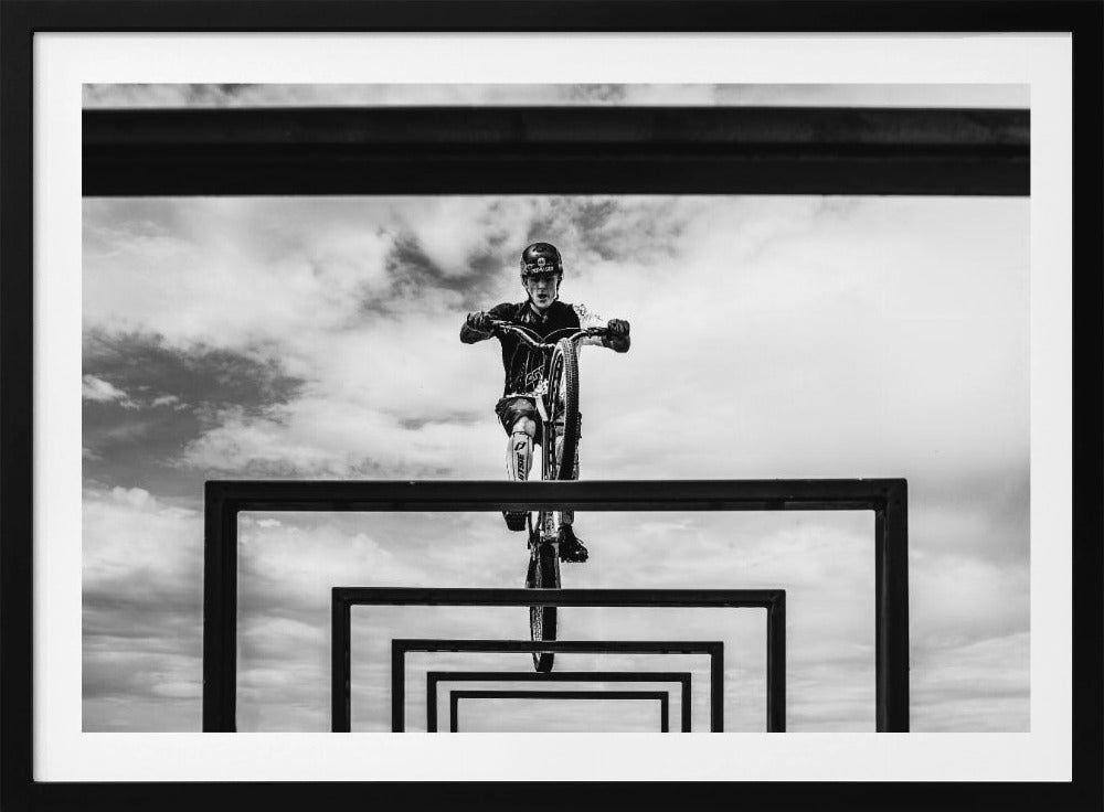 A striking black and white photograph taken from a low angle, showing a cyclist in full gear balancing on a mountain bike. The cyclist is positioned centrally above a series of black rectangular frames that create a receding, tunnel-like visual effect against a cloudy sky. The entire image is presented within a silver picture frame. Artwork