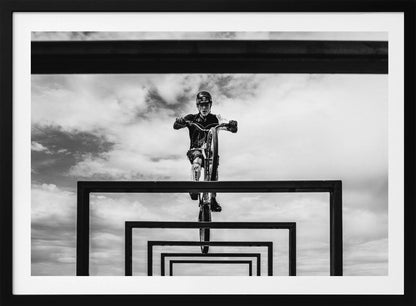 A striking black and white photograph taken from a low angle, showing a cyclist in full gear balancing on a mountain bike. The cyclist is positioned centrally above a series of black rectangular frames that create a receding, tunnel-like visual effect against a cloudy sky. The entire image is presented within a silver picture frame. Artwork