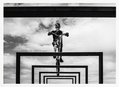 A striking black and white photograph taken from a low angle, showing a cyclist in full gear balancing on a mountain bike. The cyclist is positioned centrally above a series of black rectangular frames that create a receding, tunnel-like visual effect against a cloudy sky. The entire image is presented within a silver picture frame. Artwork