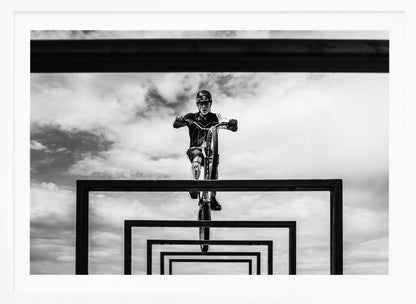 A striking black and white photograph taken from a low angle, showing a cyclist in full gear balancing on a mountain bike. The cyclist is positioned centrally above a series of black rectangular frames that create a receding, tunnel-like visual effect against a cloudy sky. The entire image is presented within a silver picture frame. Artwork