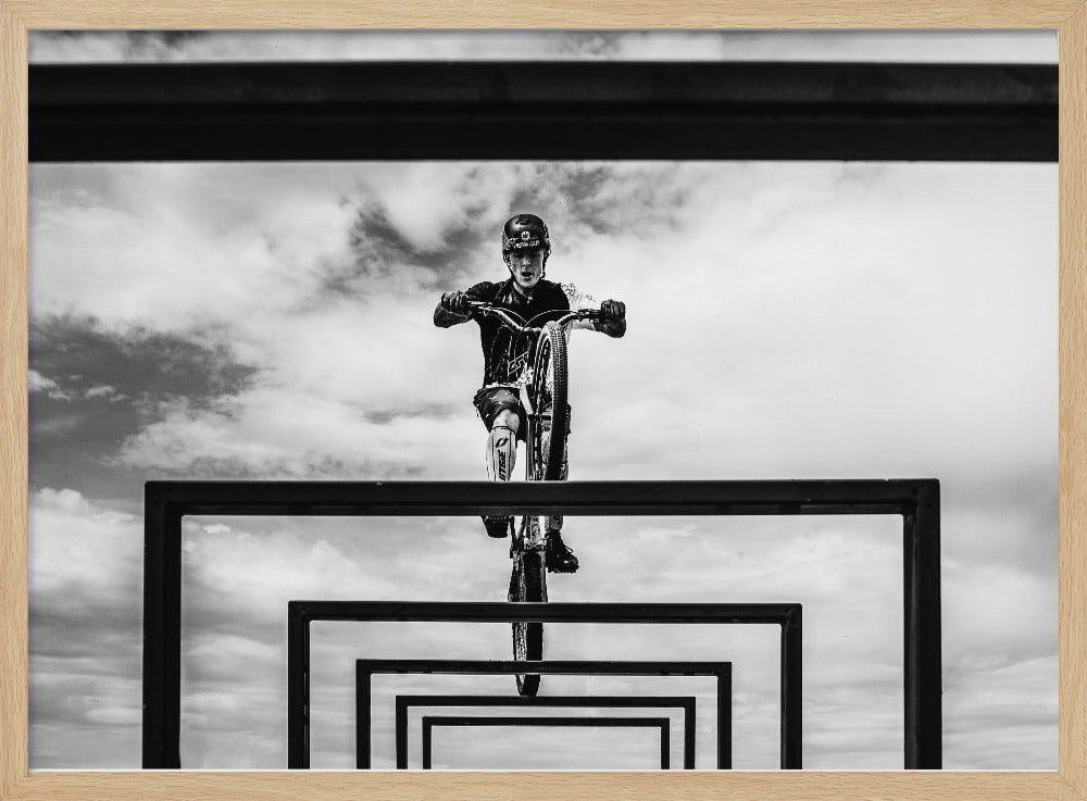 A striking black and white photograph taken from a low angle, showing a cyclist in full gear balancing on a mountain bike. The cyclist is positioned centrally above a series of black rectangular frames that create a receding, tunnel-like visual effect against a cloudy sky. The entire image is presented within a silver picture frame. Artwork