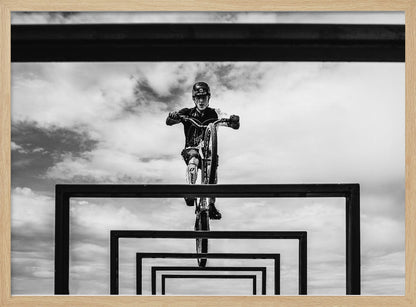 A striking black and white photograph taken from a low angle, showing a cyclist in full gear balancing on a mountain bike. The cyclist is positioned centrally above a series of black rectangular frames that create a receding, tunnel-like visual effect against a cloudy sky. The entire image is presented within a silver picture frame. Artwork