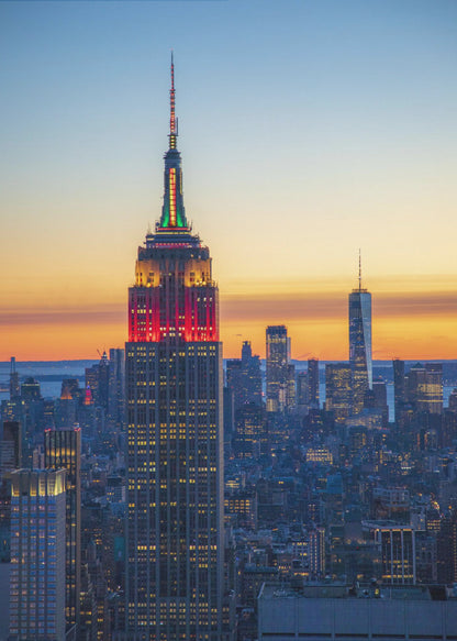 A vertical photograph of the New York City skyline at sunset, with the Empire State Building in the foreground. The building is illuminated with festive red and green lights on its upper floors, set against a sky that transitions from deep blue to a warm orange glow at the horizon. The surrounding cityscape, including the distant One World Trade Center, is bathed in the cool light of dusk, with scattered warm lights from building windows. Decor